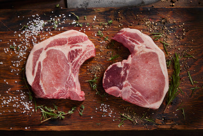 Two raw pork chops on a wooden cutting board with herbs and salt.