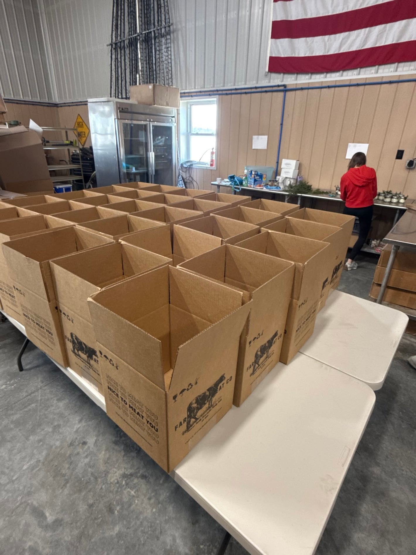 Multiple open brown cardboard shipping boxes arranged on white tables in a warehouse or processing space, with a person visible in the background near a worktable and an American flag hanging on the wall.