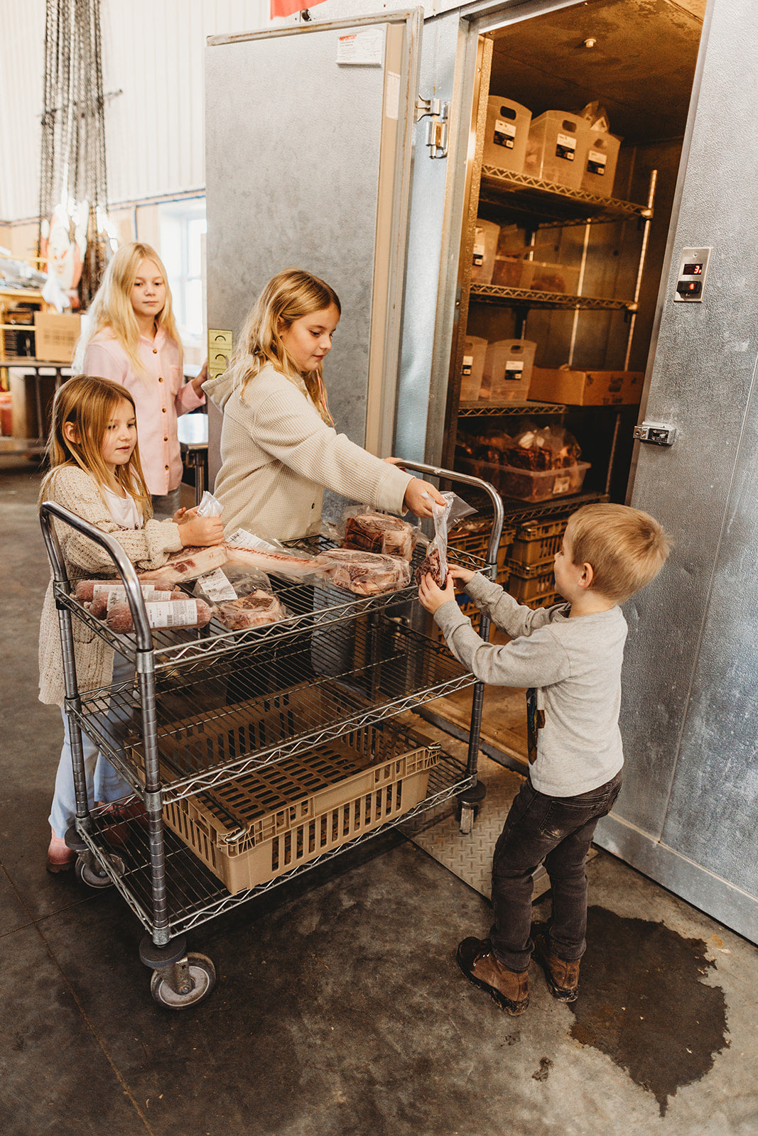 Children unload packaged goods from a large metal utility cart positioned in front of an open industrial storage room door with shelves of boxes.