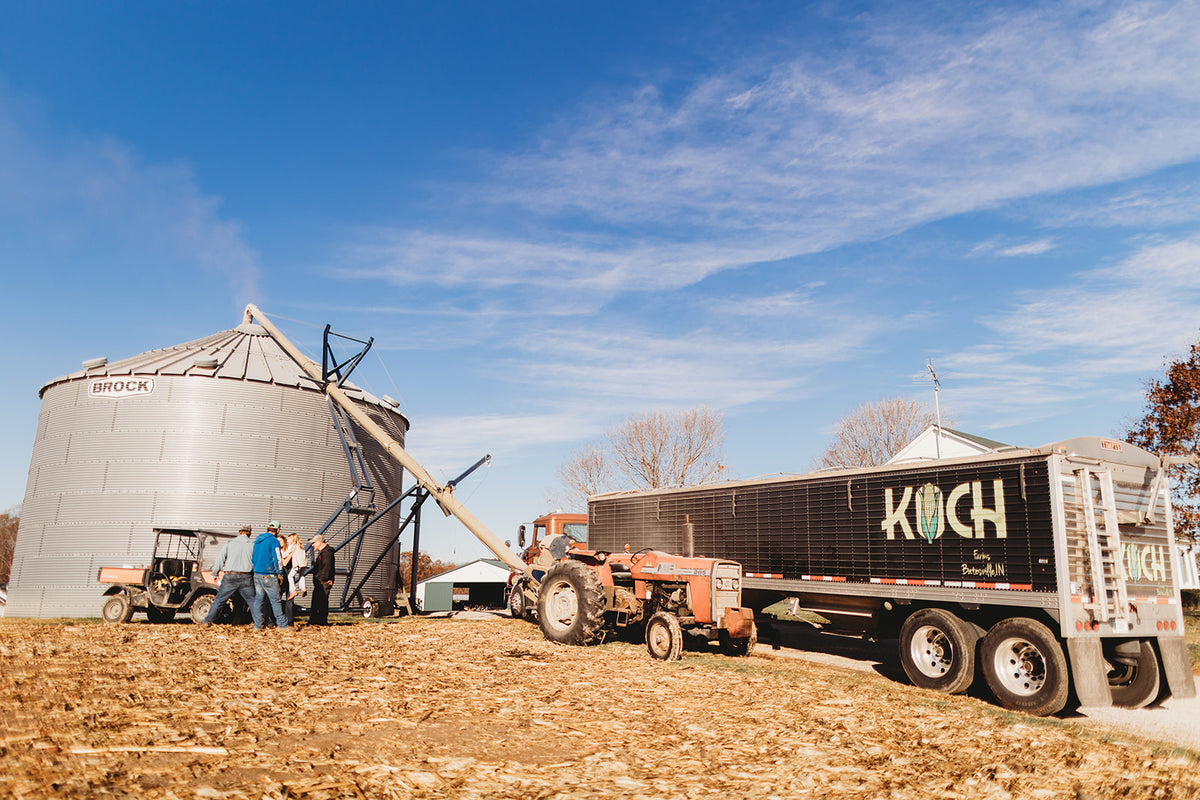 Tractor and trailer with 'Koch' branding on a farm with silos and blue sky.
