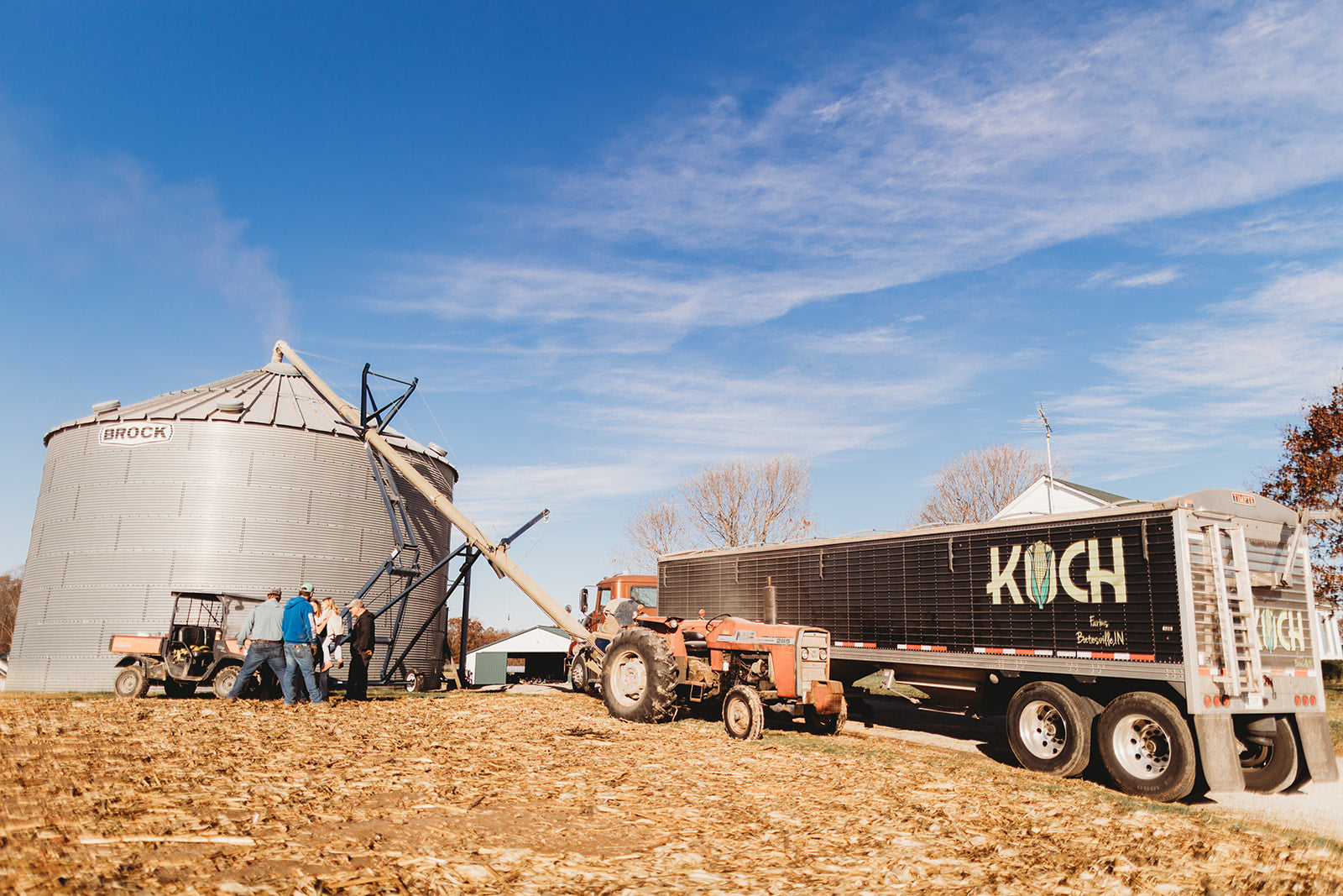 Tractor and trailer with 'Koch' branding on a farm with silos and blue sky.