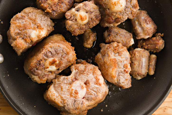 Browned oxtail pieces in a black pan on a wooden surface