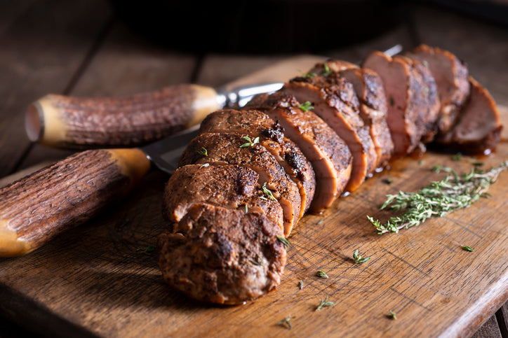 Sliced meat on a wooden board with a knife, garnished with herbs.